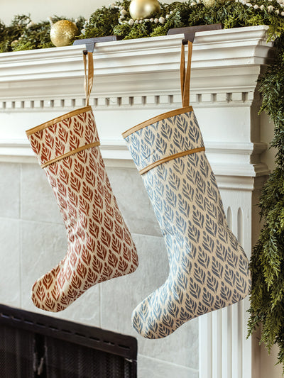 Red and blue hand-block printed Christmas stockings styled on a holiday mantel with evergreen garland, wooden beads, and gold ornaments for sustainable seasonal décor.