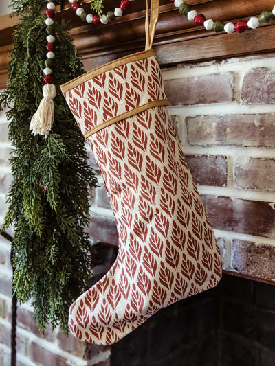 Single red hand-block printed Christmas stocking with modern leaf motif hanging on a rustic brick fireplace mantel, styled with cedar greenery and a festive wooden bead garland — ethically made holiday décor.
