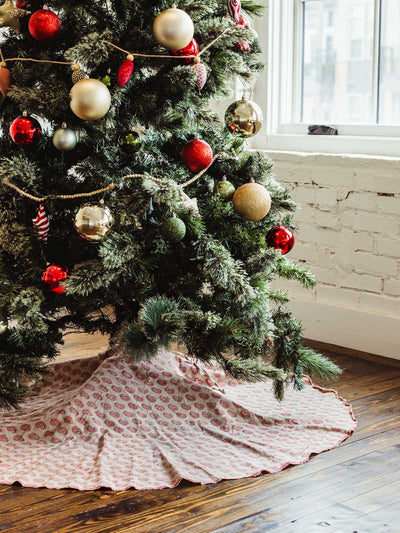 Christmas tree decorated with red, gold, and green ornaments and wooden bead garland, styled with a hand-block printed red and cream tree skirt on hardwood floors — artisanal holiday décor.