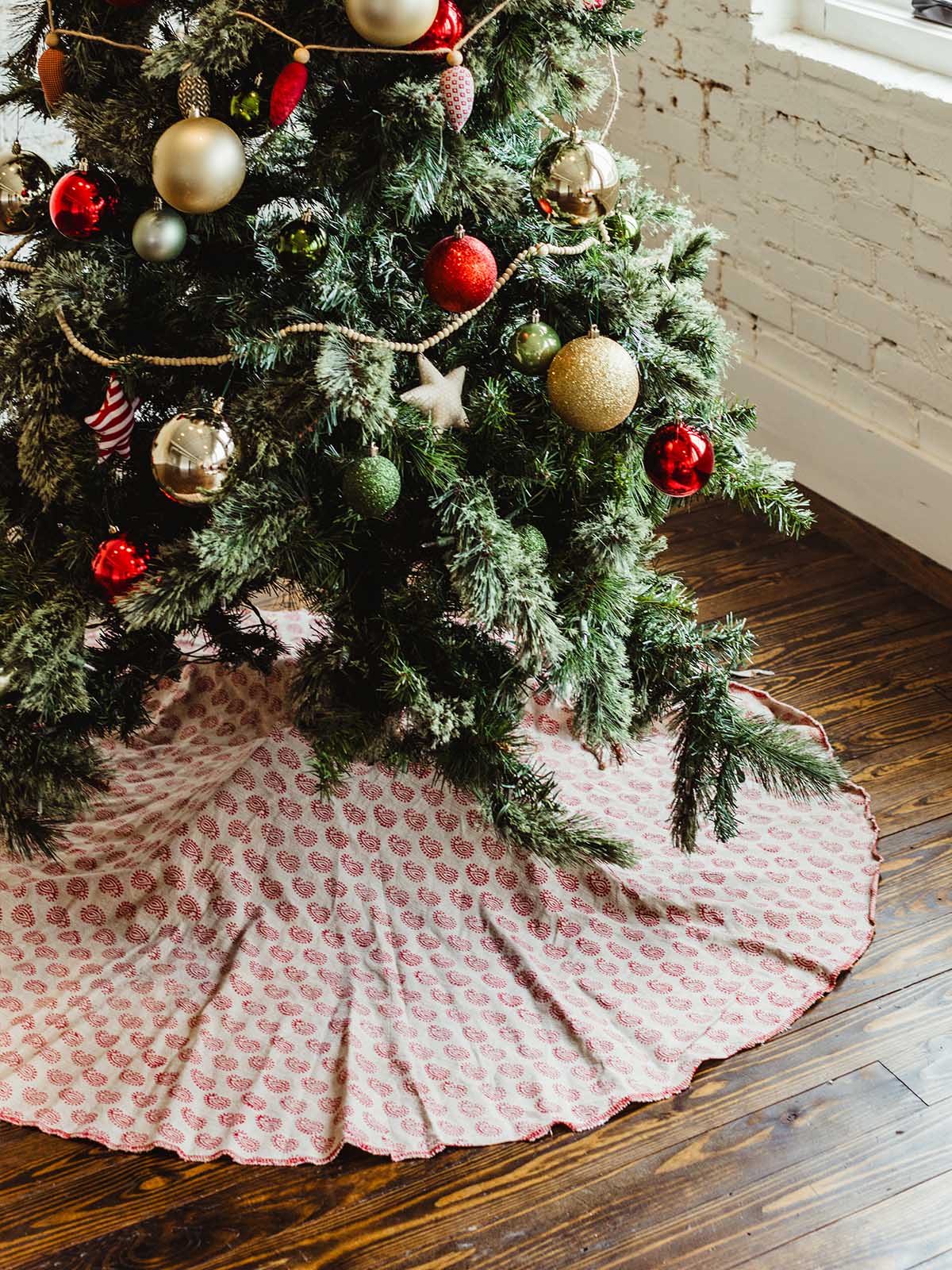 Full Christmas tree with red and gold baubles and handmade wooden bead garland, featuring a red and cream patterned block print tree skirt — ethically crafted festive home styling.