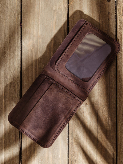 A brown leather wallet is displayed open on a wooden surface, showing multiple card slots and a transparent ID window. Sunlight and shadows create a natural, textured look across the image.