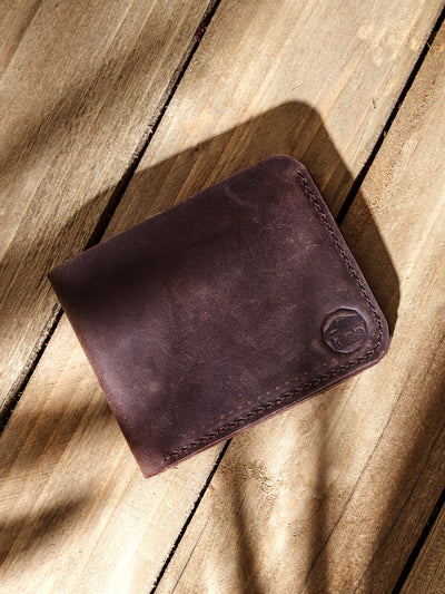 Closed dark brown leather wallet embossed with the Kilele logo in the corner, resting on a wood plank surface with natural shadowing.
