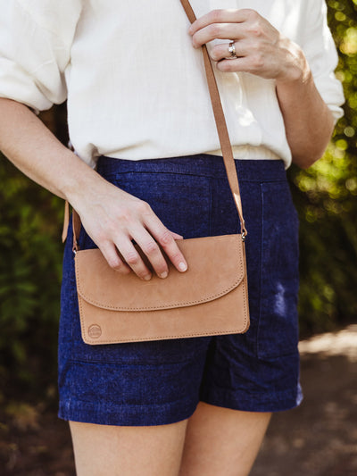 Close-up of a woman wearing a white blouse and navy shorts, holding a tan leather crossbody wallet with a flap closure and slim strap, outdoors with greenery in the background.