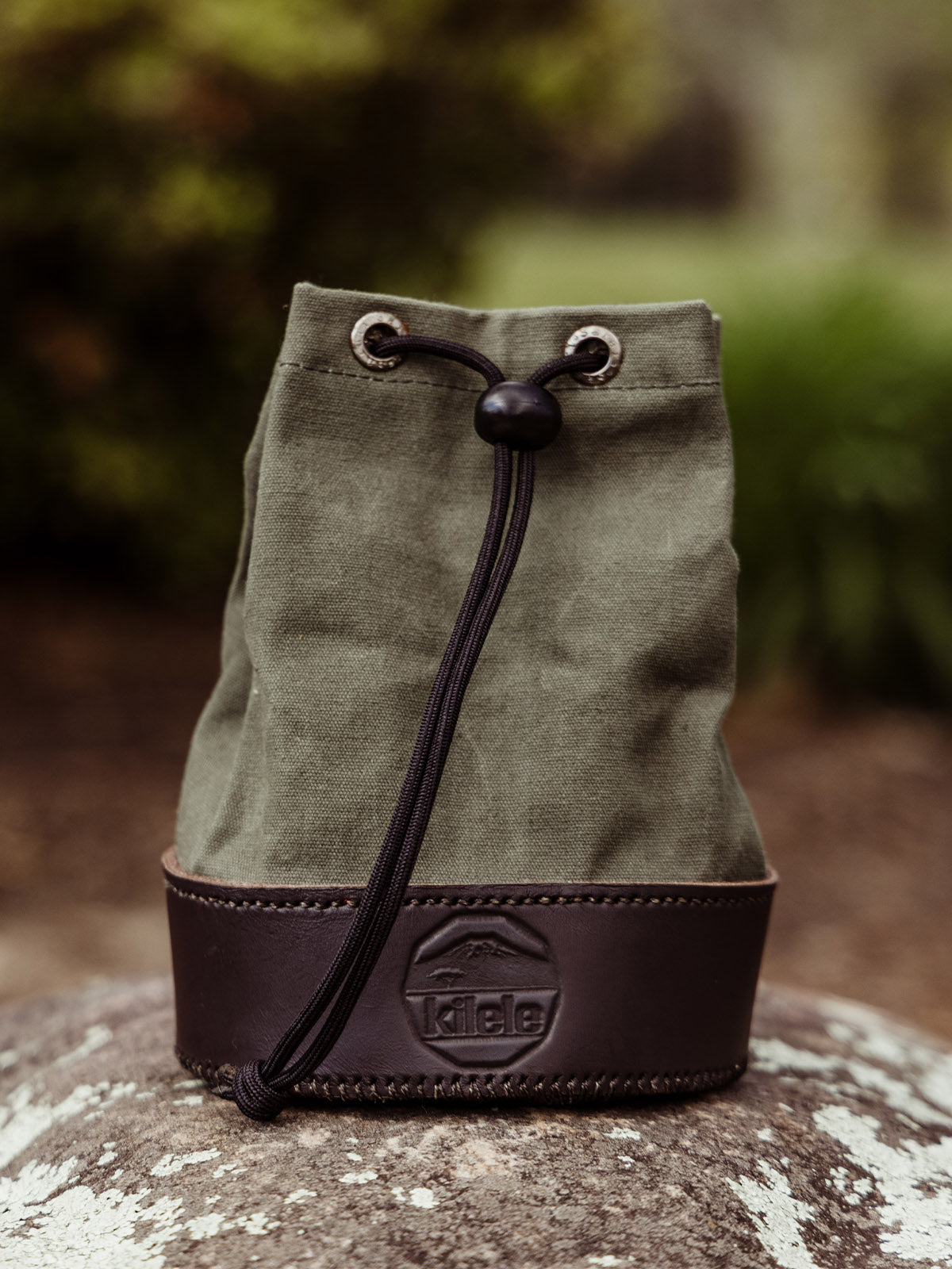  A green canvas and dark leather shell bag with a drawstring closure sits on a rock outdoors. The Kilele logo is embossed on the leather base.