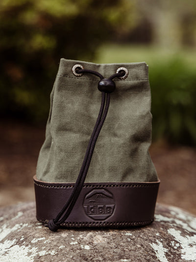  A green canvas and dark leather shell bag with a drawstring closure sits on a rock outdoors. The Kilele logo is embossed on the leather base.