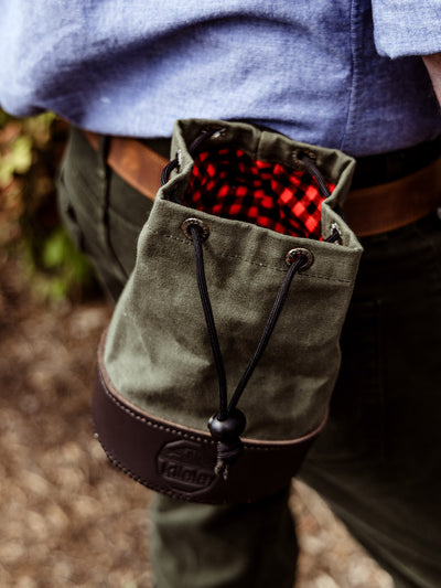 Close-up of the shell bag clipped to a man's waist, revealing a red and black plaid interior lining through the open drawstring top.