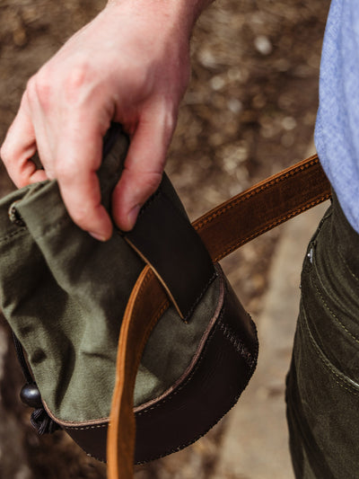 A hand is seen holding the green and leather shell bag while threading a brown leather belt through the back loop, demonstrating how it attaches.
