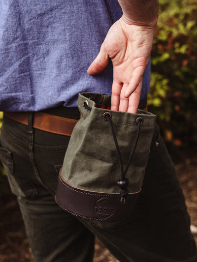 A man wearing a blue shirt and dark green pants reaches into the open shell bag clipped to his waist, showing its accessibility and practical design.