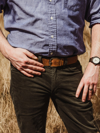A man stands with one hand on his hip and the other by his side, wearing a brown leather belt, dark pants, and a blue button-up shirt. The background shows tall, golden grass in a field.