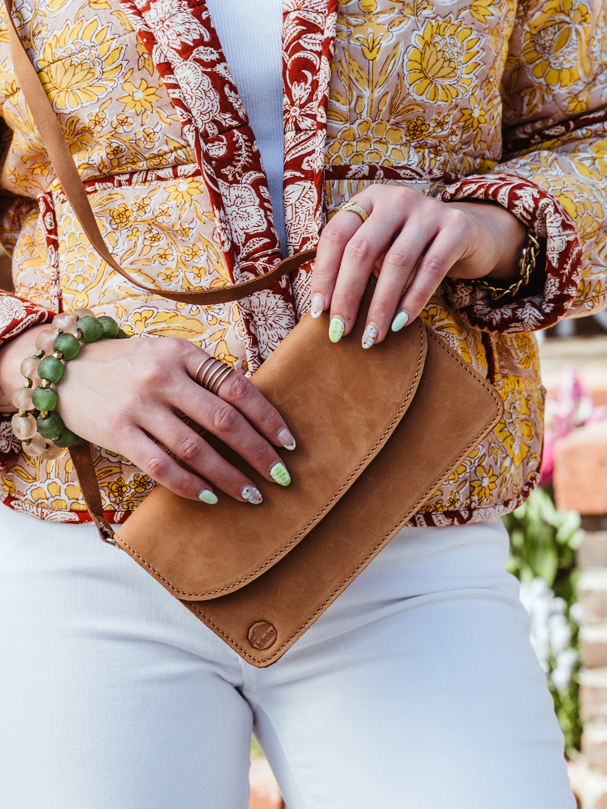 A close-up of a woman holding a tan leather crossbody wallet against her waist. She wears a colorful floral quilted jacket, white jeans, and several rings and chunky bracelets. Her nails are painted with green and white abstract designs.
