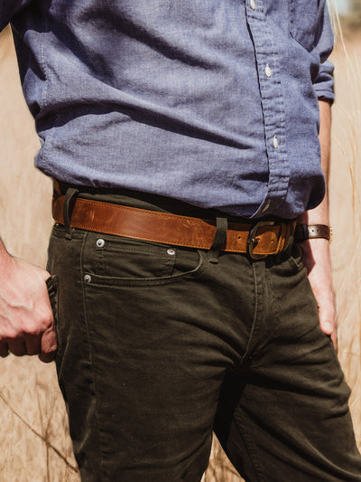  A man stands in a field wearing a light blue button-up shirt tucked into dark olive pants, showcasing a rich brown leather belt with a black trim detail.