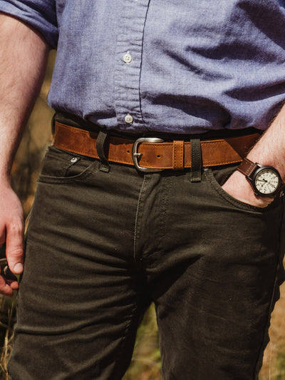 Close-up of a man’s waist wearing a light blue button-up shirt, dark pants, and a brown leather belt with a silver buckle. His left hand rests in his pocket, showing a brown leather watch with a white face.