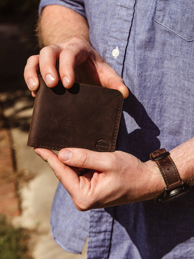 A man wearing a light blue button-up shirt and brown leather watch holds a dark brown leather wallet with a stamped logo that reads "Kilele." The photo is taken outdoors in natural light.