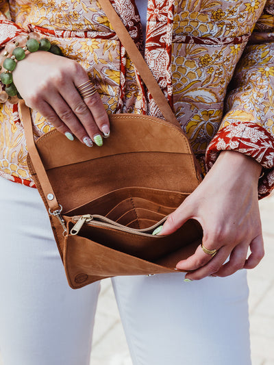 Close-up of a woman opening a tan leather crossbody wallet with multiple card slots and a zippered pocket. She wears a colorful floral quilted jacket, white pants, green and pink beaded bracelets, and a gold multi-band ring.