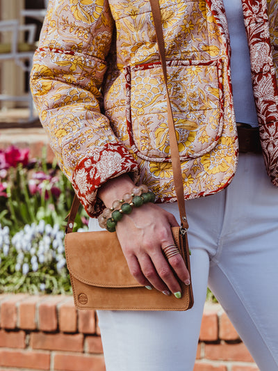 A woman standing outdoors holding a closed tan leather crossbody wallet with a button flap. She wears a floral quilted jacket in red, yellow, and cream tones, white pants, and statement bracelets.