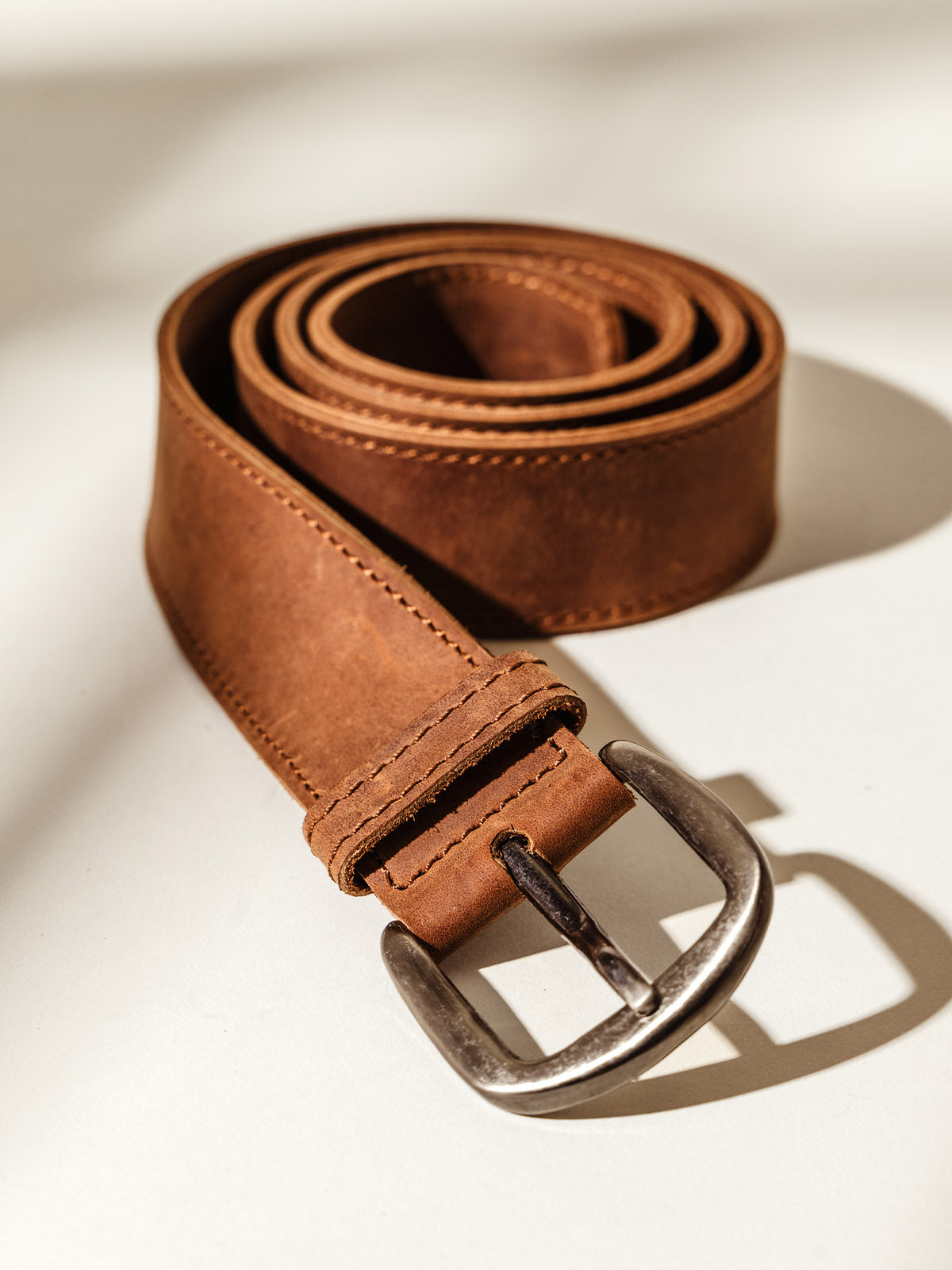 Rolled brown leather belt with visible stitching and a silver buckle, displayed on a neutral background with soft shadowing.