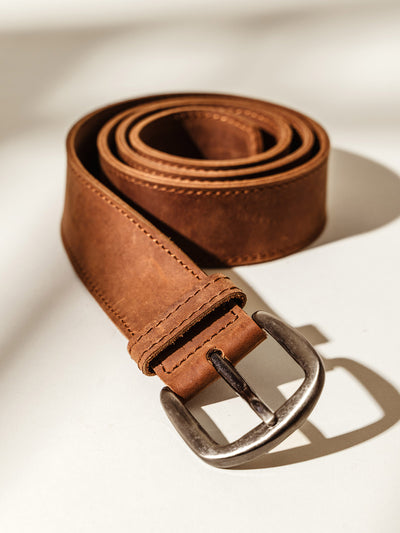 Rolled brown leather belt with visible stitching and a silver buckle, displayed on a neutral background with soft shadowing.