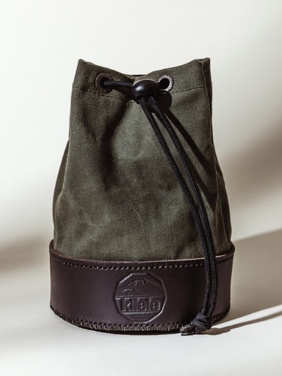 Olive green canvas shell bag with a dark leather base and drawstring closure, featuring the embossed "Kilele" logo, styled against a light neutral background.