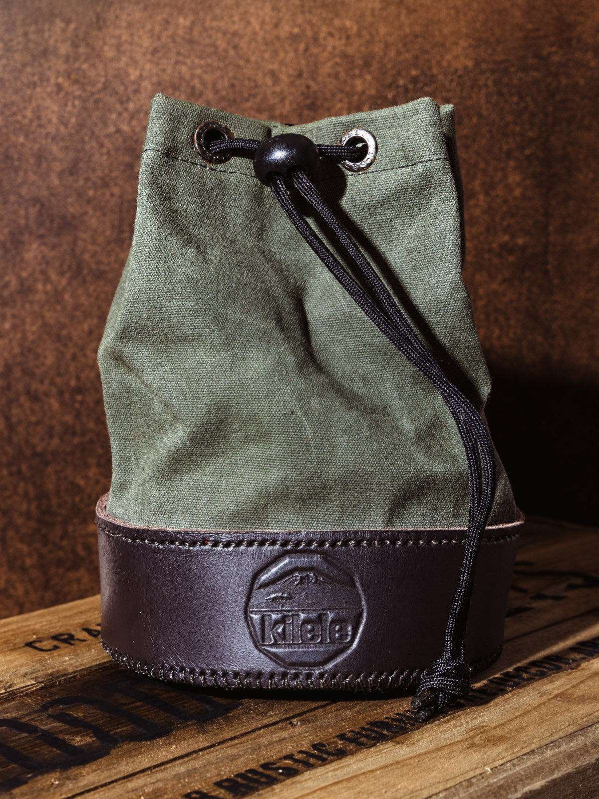 Side angle of the olive canvas and black leather Kilele shell bag with drawstring closure, displayed on a wood crate to showcase the structured design and texture.