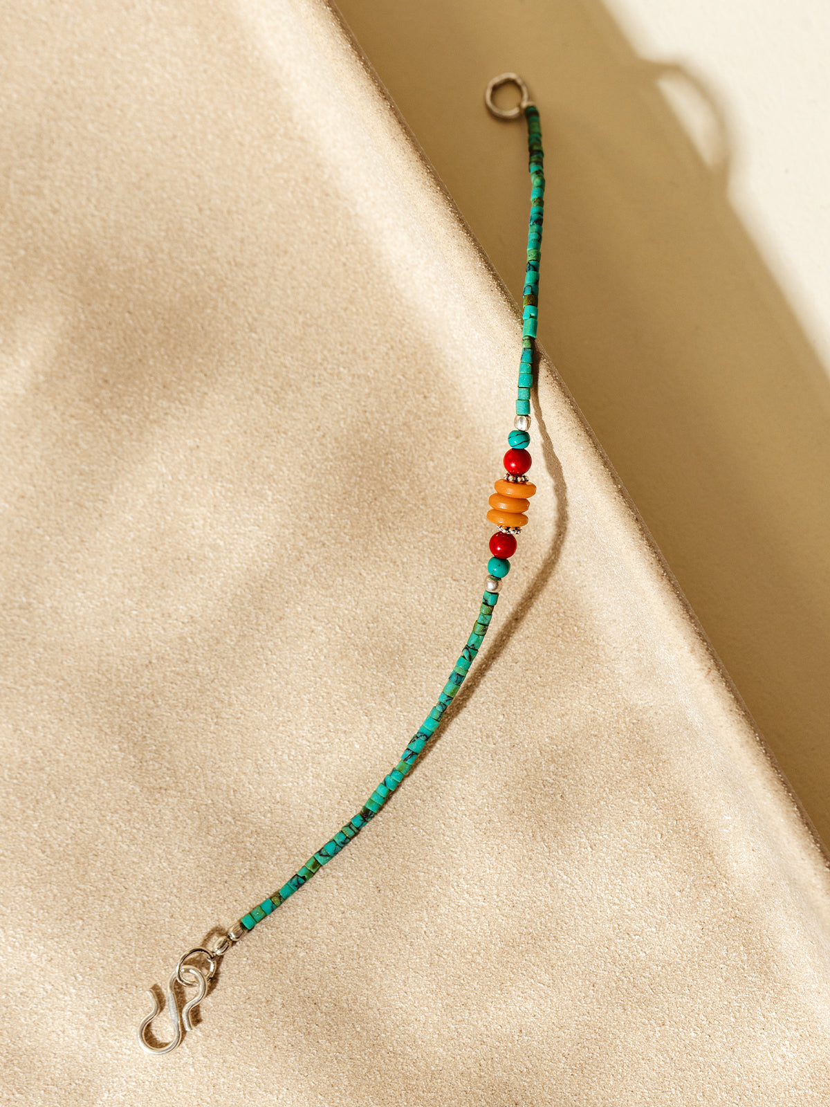 Handcrafted bracelet featuring turquoise-green beads and vibrant orange and red accents, styled on a warm beige surface with natural shadows.