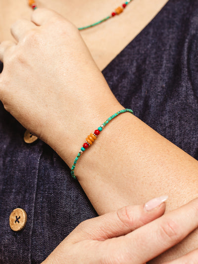 Delicate green beaded bracelet with small orange and red accent beads worn on the wrist, paired with a dark blue buttoned dress.