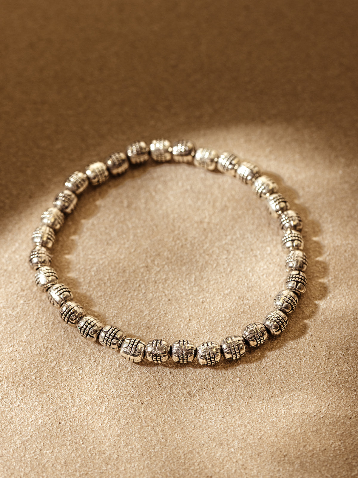 Handcrafted silver beaded bracelet arranged in a circle on a warm, neutral textured background with soft natural light highlighting the intricate bead design.