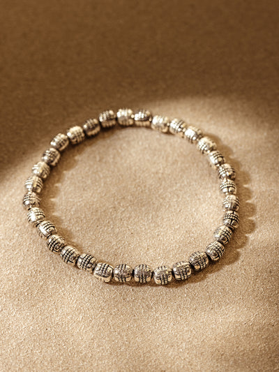 Handcrafted silver beaded bracelet arranged in a circle on a warm, neutral textured background with soft natural light highlighting the intricate bead design.