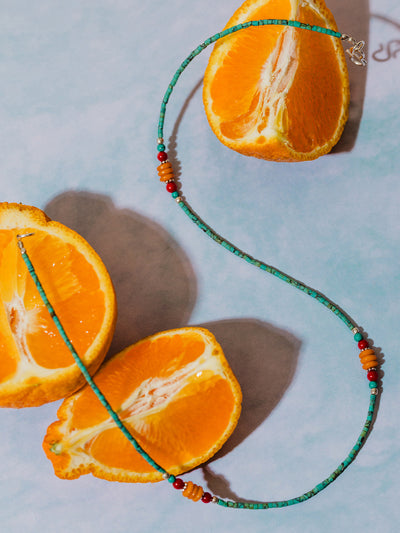 Handmade beaded necklace with turquoise, red, and amber-colored beads styled on a pastel surface with halved oranges, evoking a vibrant and fresh aesthetic.