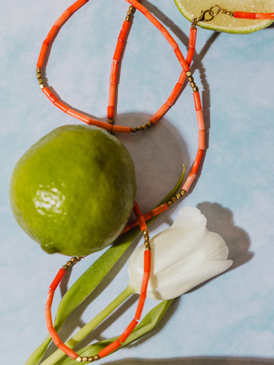Flat lay of a coral and brass beaded necklace styled with a lime, lime slice, and white tulip on a soft blue background.