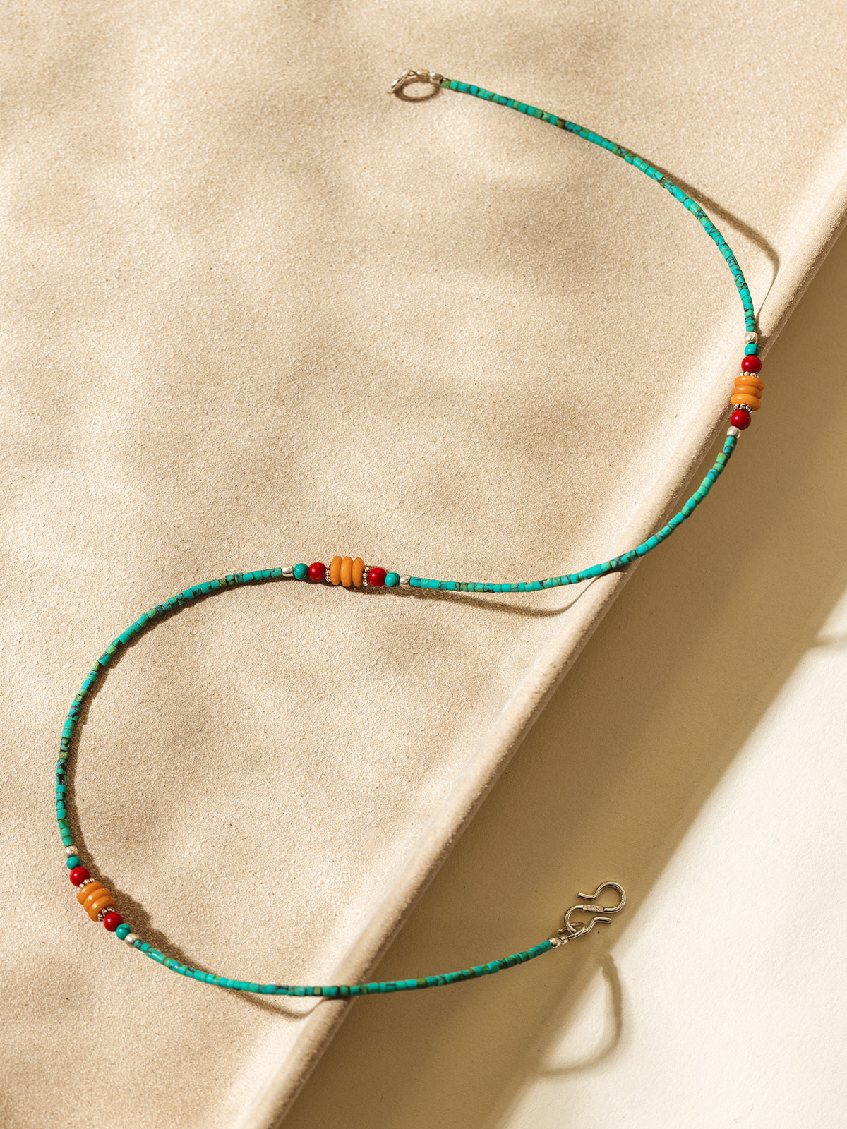 Flat lay of a colorful beaded necklace featuring turquoise, red, and amber beads arranged in an S-shape on a sunlit neutral textured surface.
