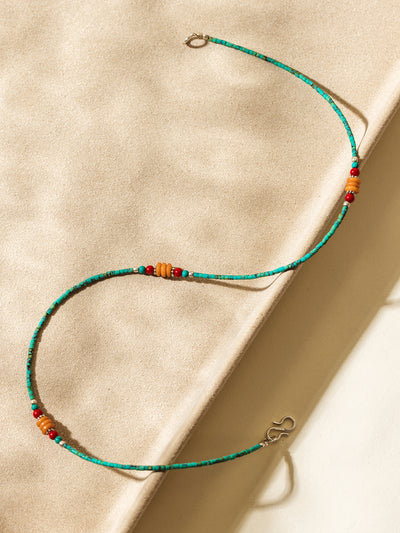 Flat lay of a colorful beaded necklace featuring turquoise, red, and amber beads arranged in an S-shape on a sunlit neutral textured surface.