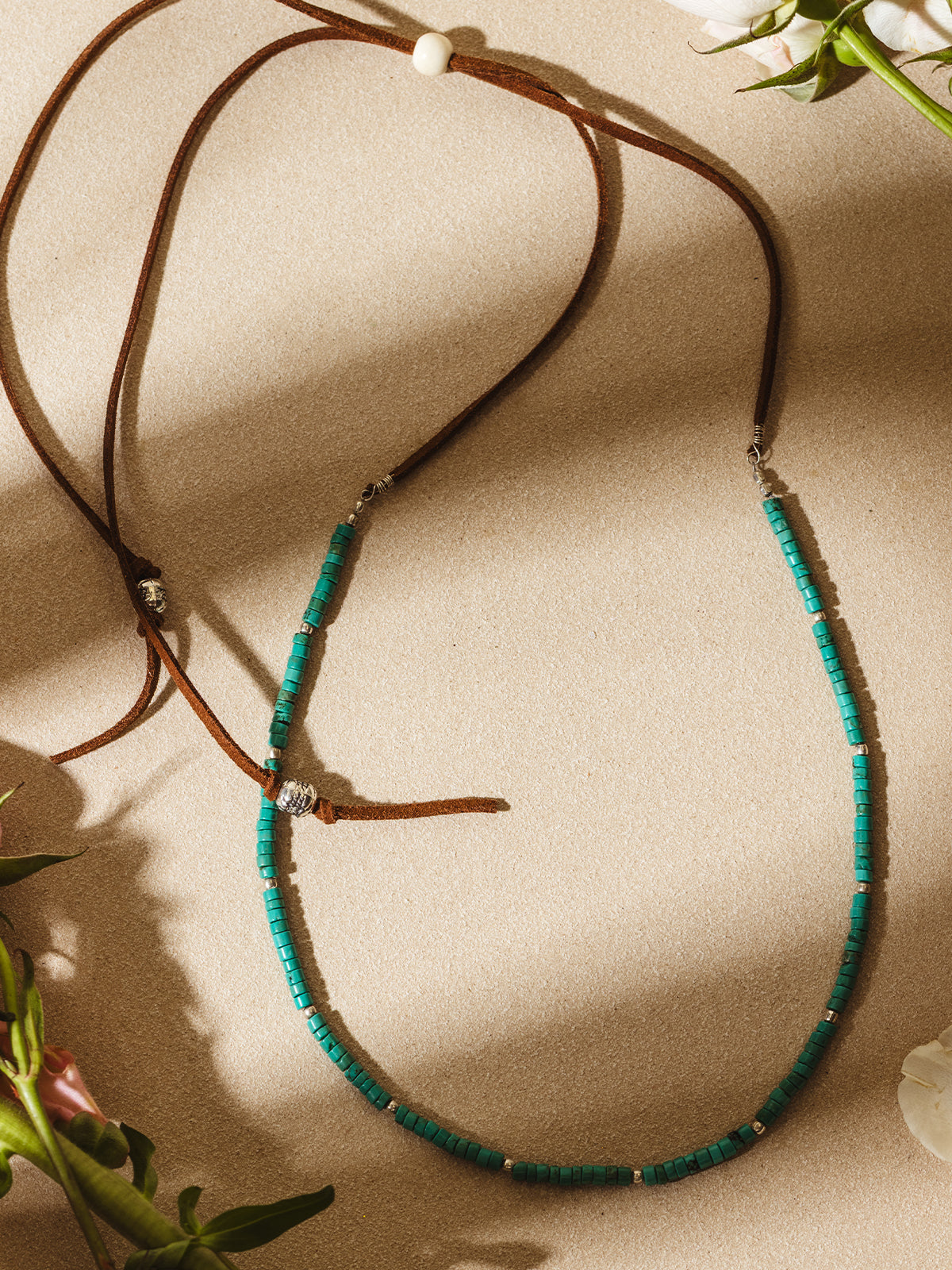 Flat lay of a green beaded necklace with silver accents and brown suede cord, styled with soft shadows and floral accents on a beige background.