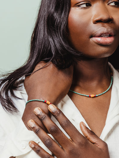 Minimalist green beaded bracelet with orange and red accent beads worn with a coordinating necklace, styled on a woman in a white button-up shirt.