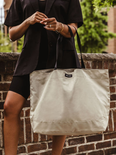 A close-up of a woman in a black blazer and bike shorts holding a large, beige canvas tote bag with black straps, standing against a brick wall.