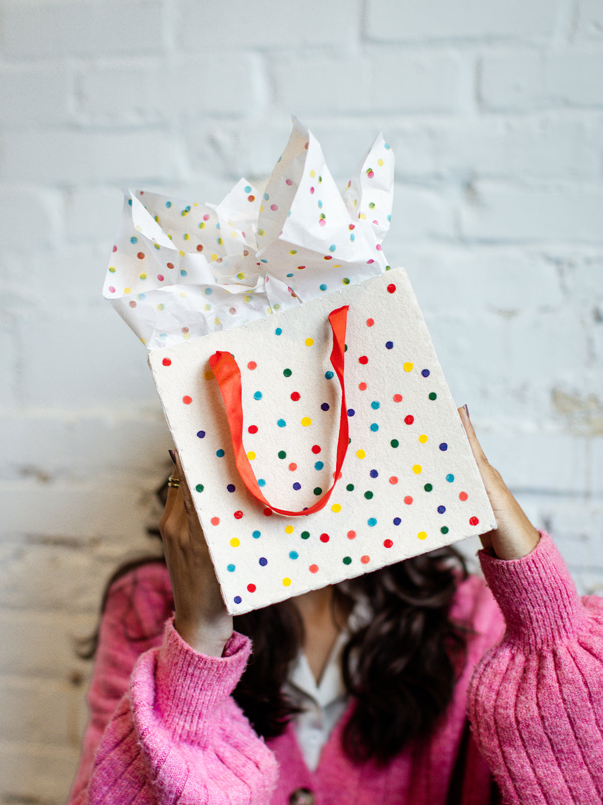 Colorful polka dot gift bag with red ribbon handles and matching tissue paper, held in front of a white brick wall by a woman in a pink sweater.