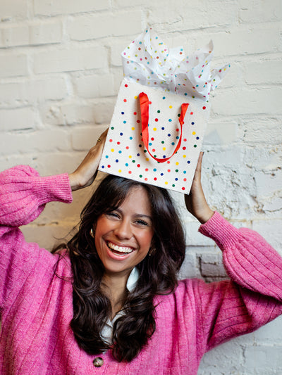 Smiling woman in a bright pink cardigan holding a multicolored polka dot gift bag with red handles and tissue paper on her head against a white brick wall.