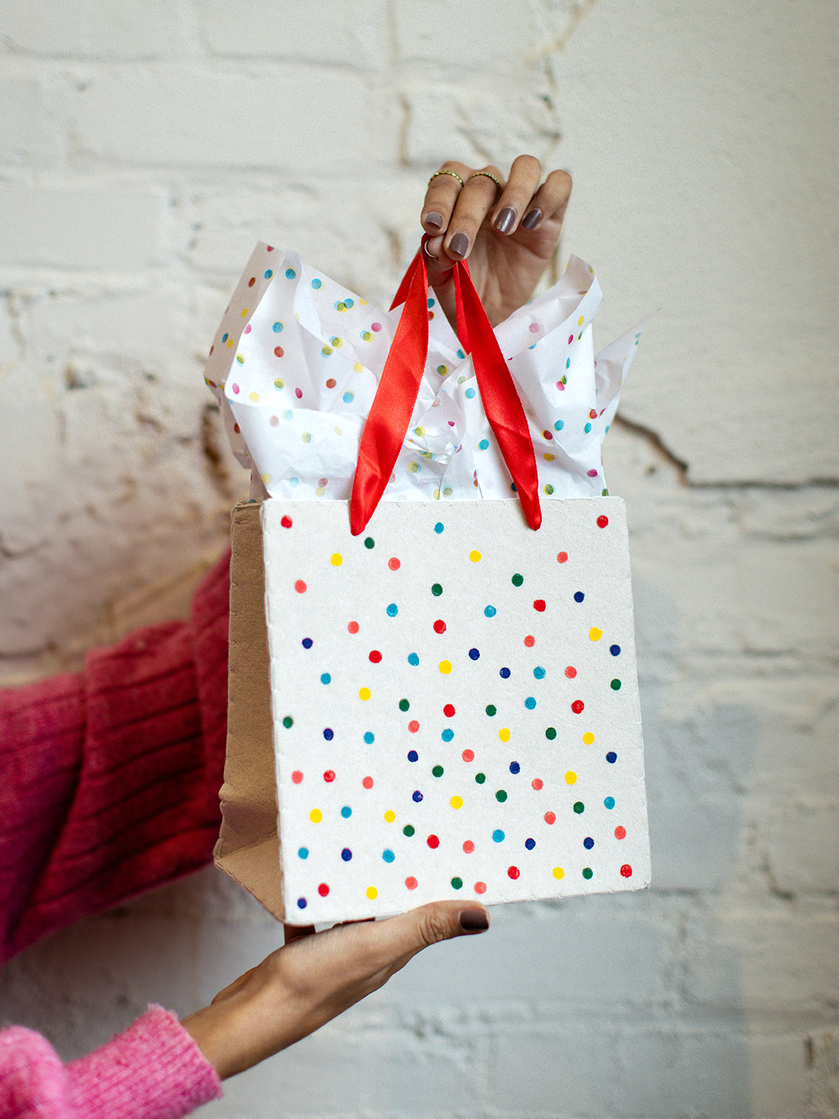 Hand holding eco-friendly white gift bag with colorful polka dots, red ribbon handles, and tissue paper, styled against a textured white brick wall.
