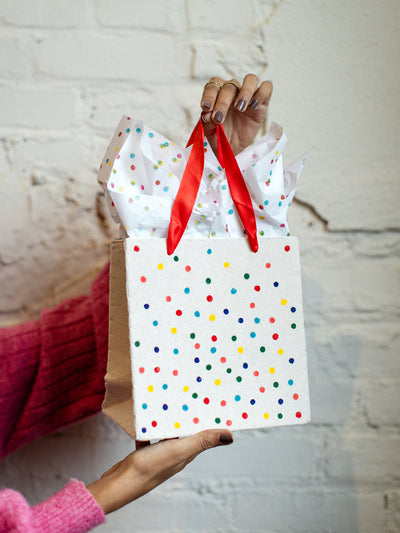 Hand holding eco-friendly white gift bag with colorful polka dots, red ribbon handles, and tissue paper, styled against a textured white brick wall.