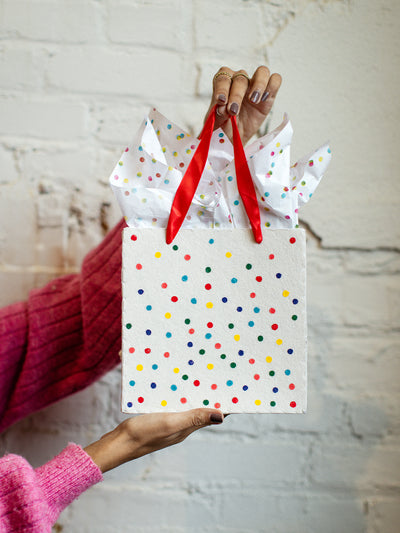 Close-up of handmade polka dot gift bag with red handles and tissue paper, held by a woman wearing a pink sweater and gold rings.