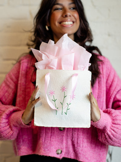 Eco-friendly handmade gift bag with embroidered pink flowers and satin ribbon handles, filled with pink tissue paper, held by a smiling woman in a pink sweater.