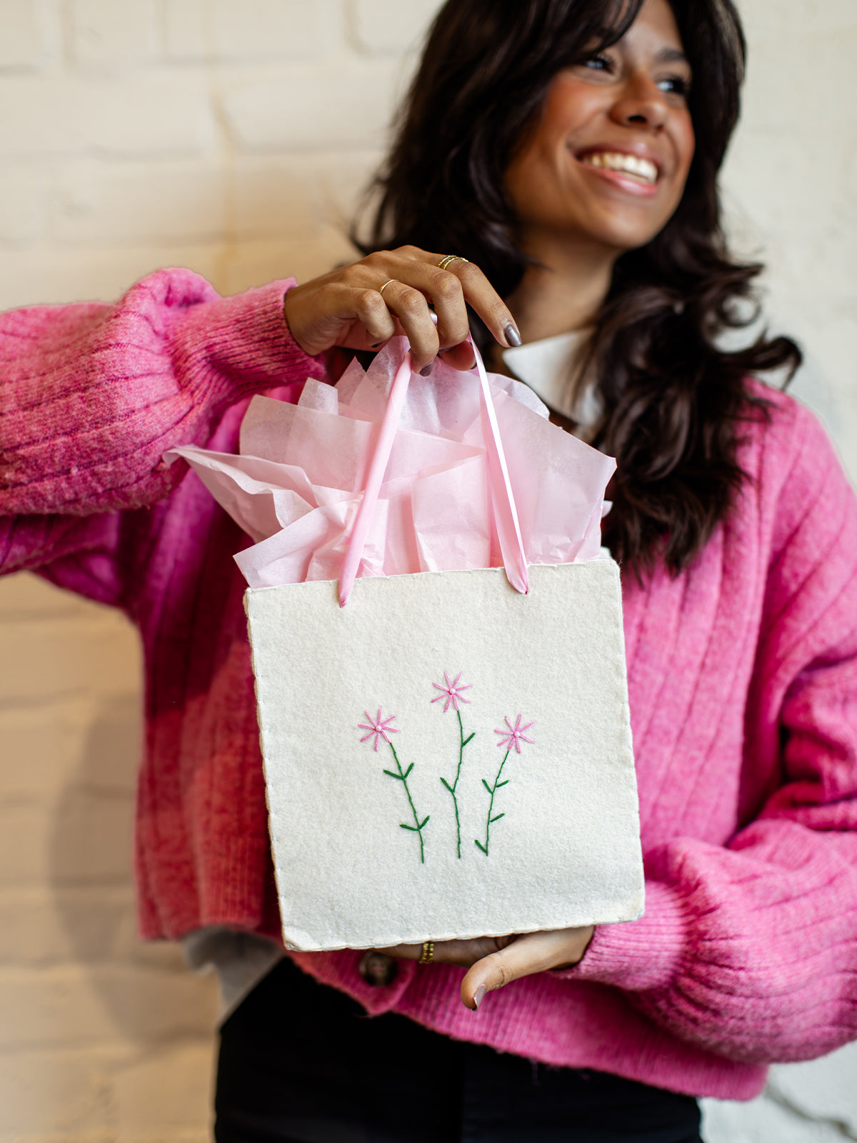 Sustainable white gift bag with pink floral embroidery and soft pink ribbon handles, styled with pink tissue paper and held by a woman in a bright pink cardigan.