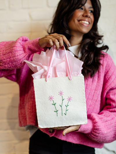 Sustainable white gift bag with pink floral embroidery and soft pink ribbon handles, styled with pink tissue paper and held by a woman in a bright pink cardigan.