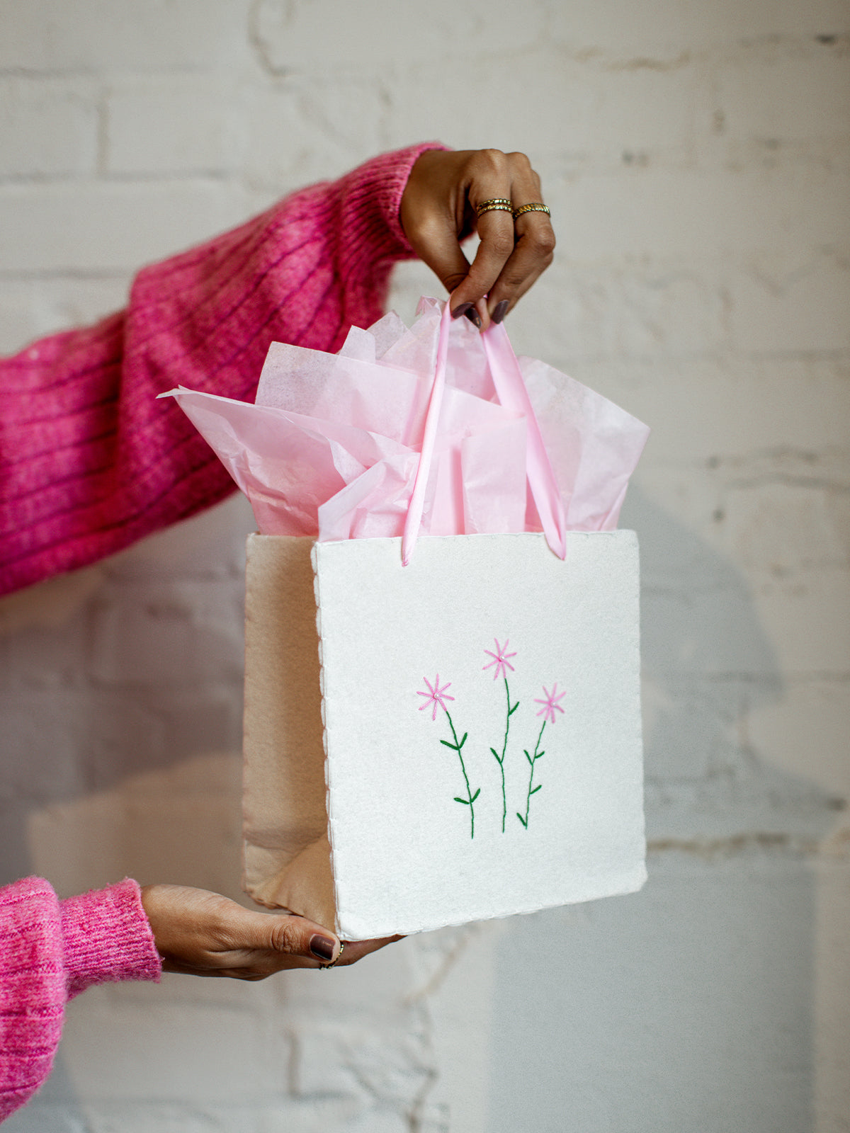 Close-up side view of handmade embroidered gift bag with pink flower details and pink handles, filled with tissue paper, held against a white brick wall.