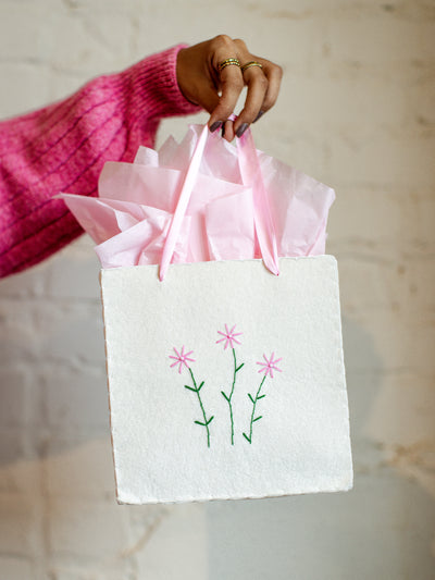 Minimalist eco-friendly gift bag featuring hand-stitched pink flowers and pink ribbon handles, displayed with pastel pink tissue paper for gifting.
