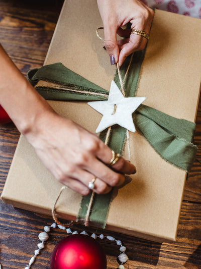 Close-up of hands tying twine on a sustainable Christmas gift box wrapped in kraft paper with green ribbon and star ornament, with red ornament nearby.