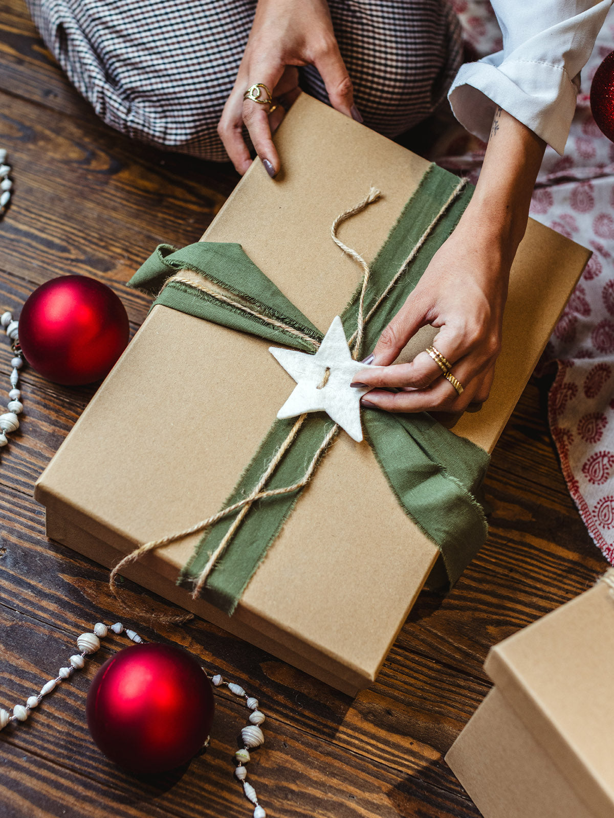 Person holding sustainable Christmas gift wrapped in kraft paper with fabric ribbon and star ornament, surrounded by festive red ornaments and holiday decor.
