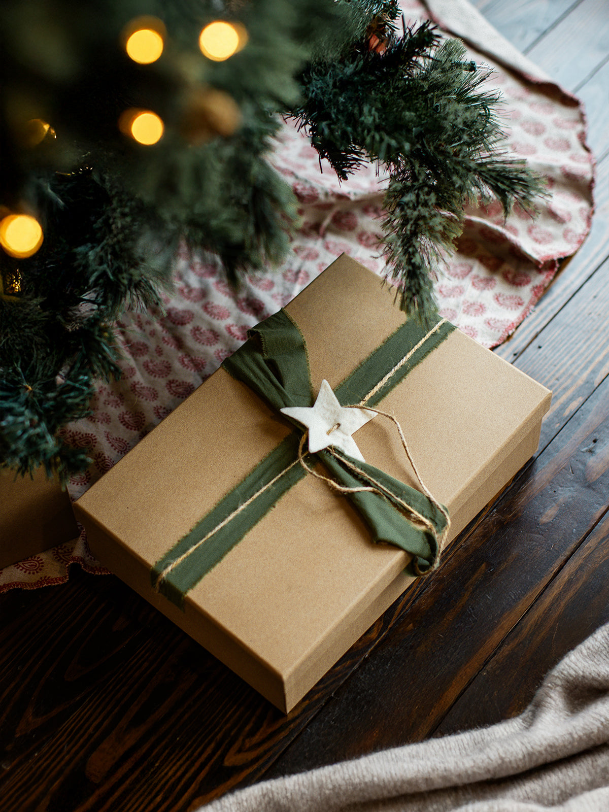 Eco-friendly holiday gift wrapped in kraft paper with green fabric ribbon and white star ornament, placed under a decorated Christmas tree.