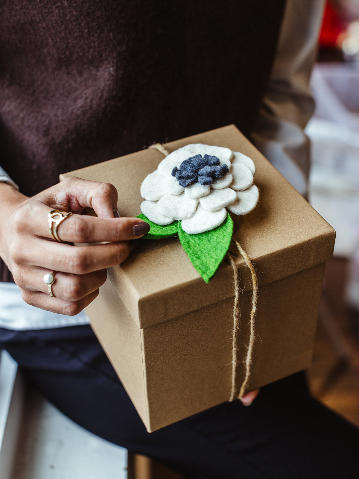 Sustainable gift box wrapped in kraft paper with twine and topped with a handmade felt flower decoration, held by a person wearing a pearl ring.