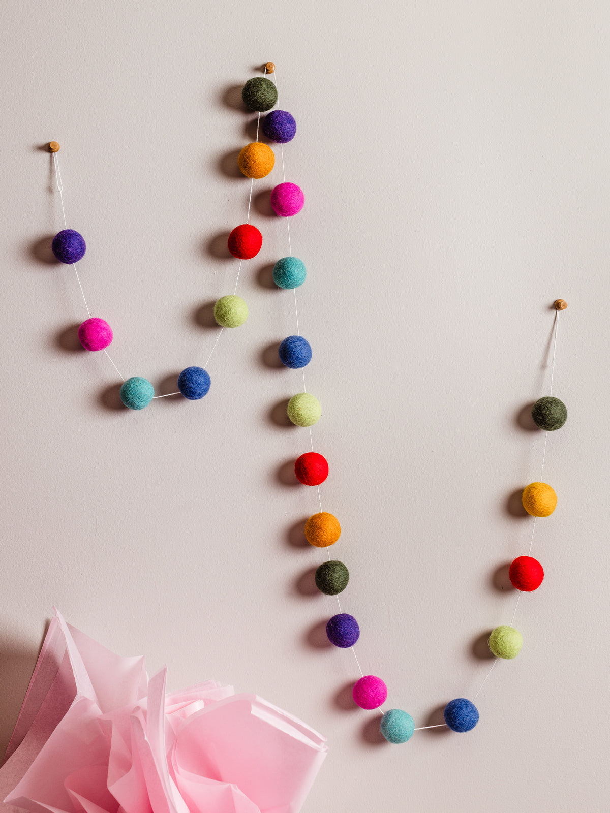Handmade wool felt ball garland in bright rainbow colors hanging on a neutral wall with pink tissue paper decor below. Sustainable birthday or party decoration.