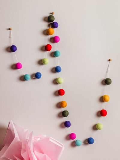 Handmade wool felt ball garland in bright rainbow colors hanging on a neutral wall with pink tissue paper decor below. Sustainable birthday or party decoration.